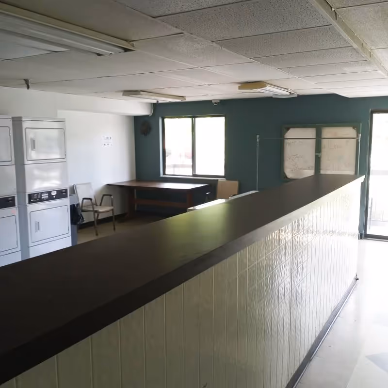 Interior common room with a long counter, stacked laundry machines, chairs and a table by windows.