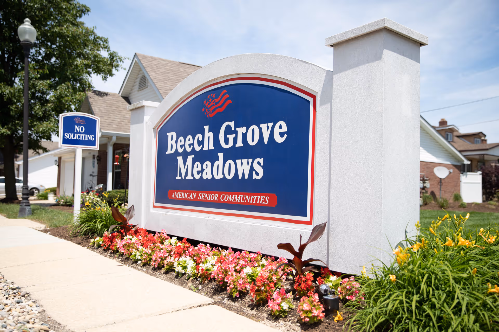 Outdoor view of a large sign for Beech Grove Meadows, an American senior community, surrounded by colorful flowers and greenery with a sidewalk and houses in the background under a clear sky.