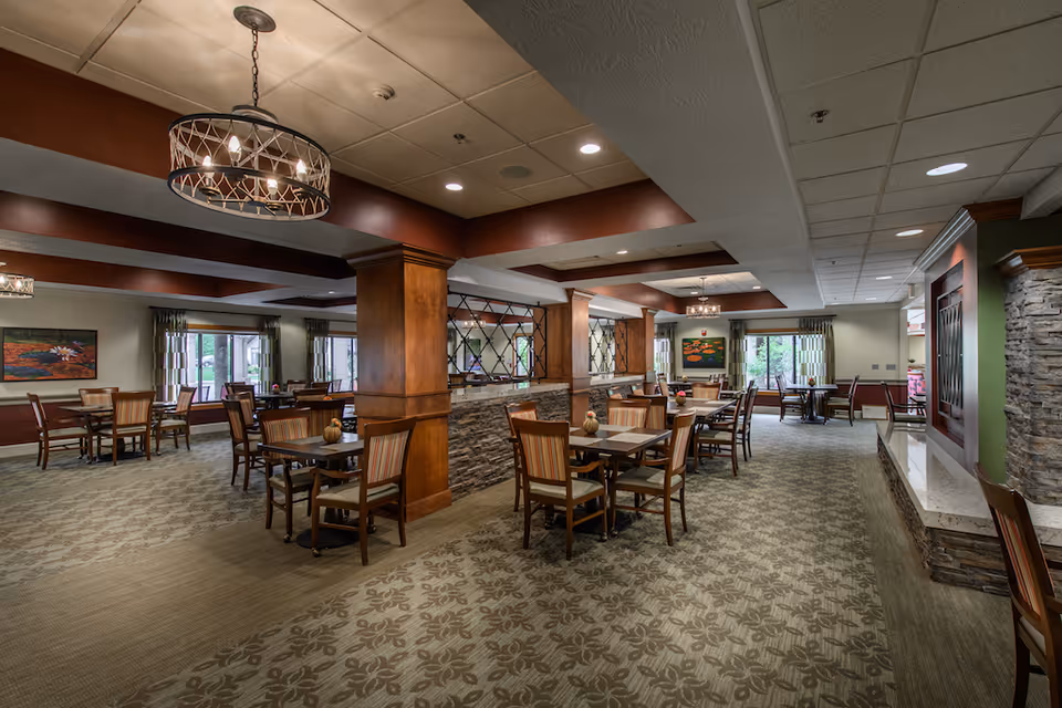 A spacious dining room in a senior living facility with multiple wooden tables and chairs arranged neatly. The room features patterned carpet, warm lighting fixtures hanging from the ceiling, large windows with curtains allowing natural light, and decorative wooden columns with stone accents.