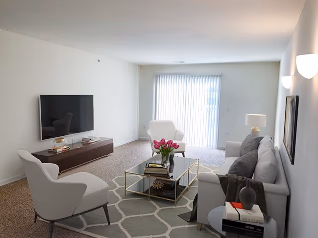 A bright living room with a large window covered by vertical blinds. The room features a gray sofa with dark cushions, a white armchair, a glass coffee table with books and a vase of pink flowers, and a wall-mounted flat-screen TV above a wooden media console. The walls are white, and the floor is carpeted with a patterned area rug.