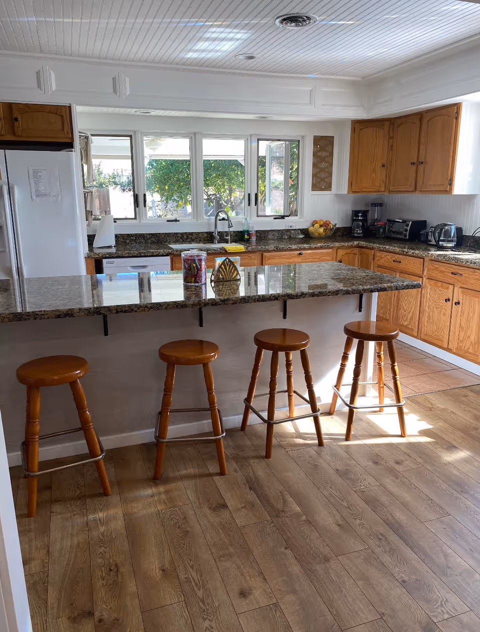 A bright kitchen with wooden cabinets, granite countertops, and a large island with four wooden stools. The kitchen features a refrigerator, sink under a window, coffee maker, toaster, and other small appliances. The floor is wood, and sunlight streams in through the windows.