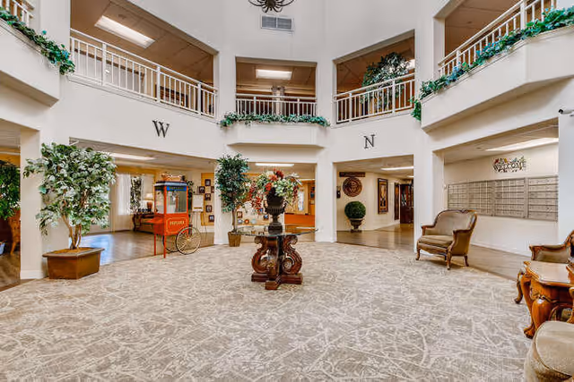Spacious and well-lit interior common area of a senior living facility with beige patterned carpet, decorative plants, a central wooden table with a floral arrangement, comfortable chairs, and a popcorn machine. The area has a high ceiling with a balcony railing on the upper level and directional letters W and N on the walls.