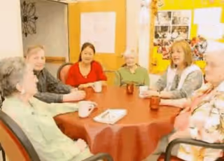 A group of six elderly women sitting around a round table covered with a red tablecloth, each holding a cup, in a cozy room with yellow walls and photos on a bulletin board in the background.