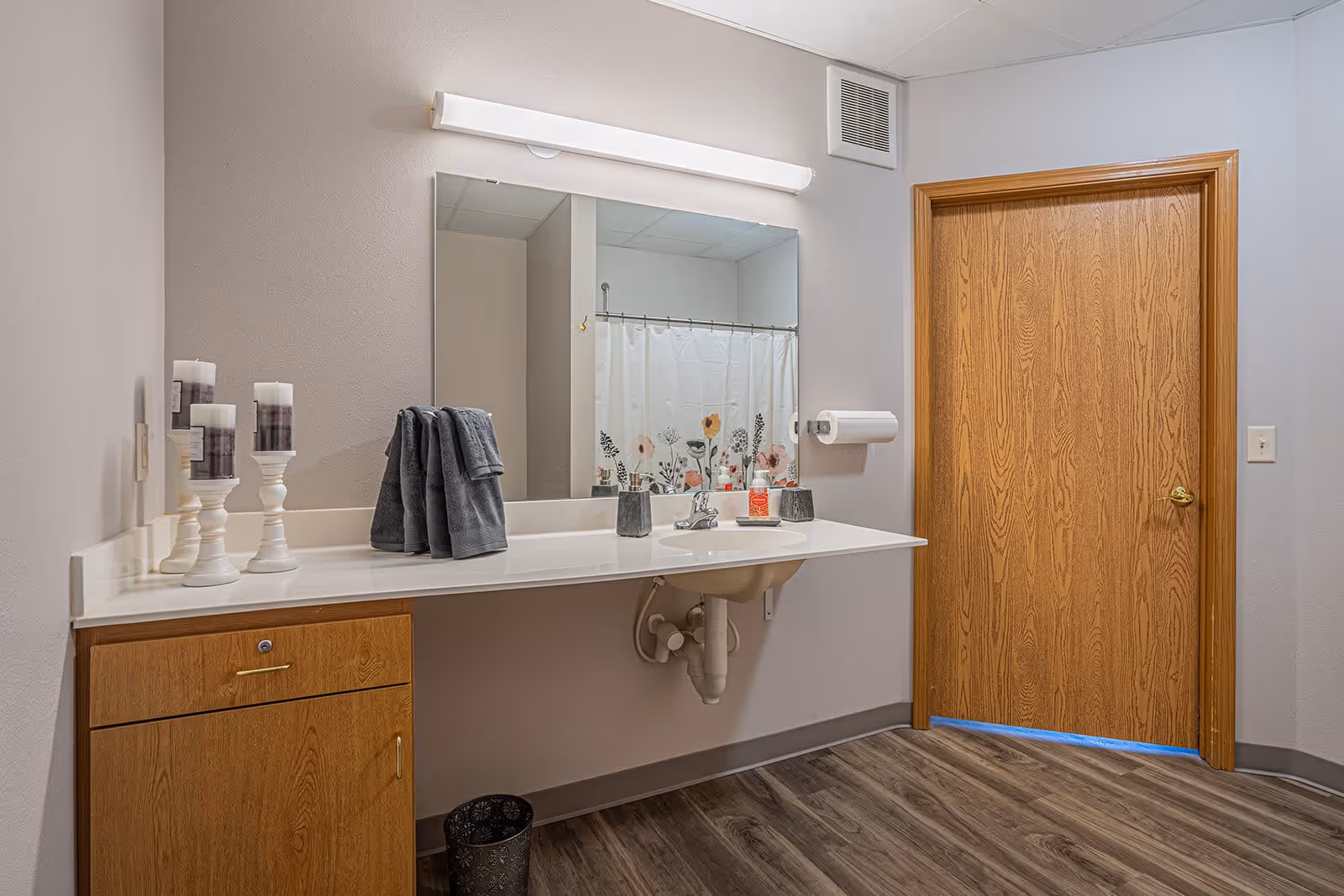 A bathroom vanity area with a large mirror above a white countertop. On the countertop are three white candle holders with candles, two folded dark gray towels, a soap dispenser, and a small container. There is a wooden cabinet below the countertop. A wooden door is visible to the right, and a shower curtain with a floral pattern is reflected in the mirror. The floor has wood-like vinyl planks.