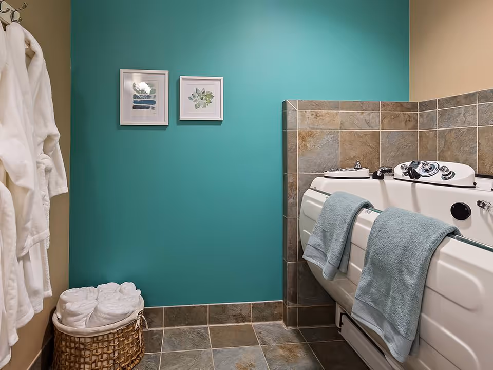 A bathroom with a walk-in bathtub featuring control knobs and two light blue towels hanging on the side. The walls are painted teal and beige, with two framed botanical prints hanging on the teal wall. There is a wicker basket filled with rolled white towels on the tiled floor, and two white bathrobes hanging on hooks on the left wall.