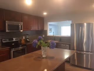 Modern kitchen with stainless steel appliances including a refrigerator, microwave, and stove. The kitchen features wooden cabinets, a brown countertop island with a vase of purple flowers, and a tiled backsplash. There is a window opening to an adjacent room.