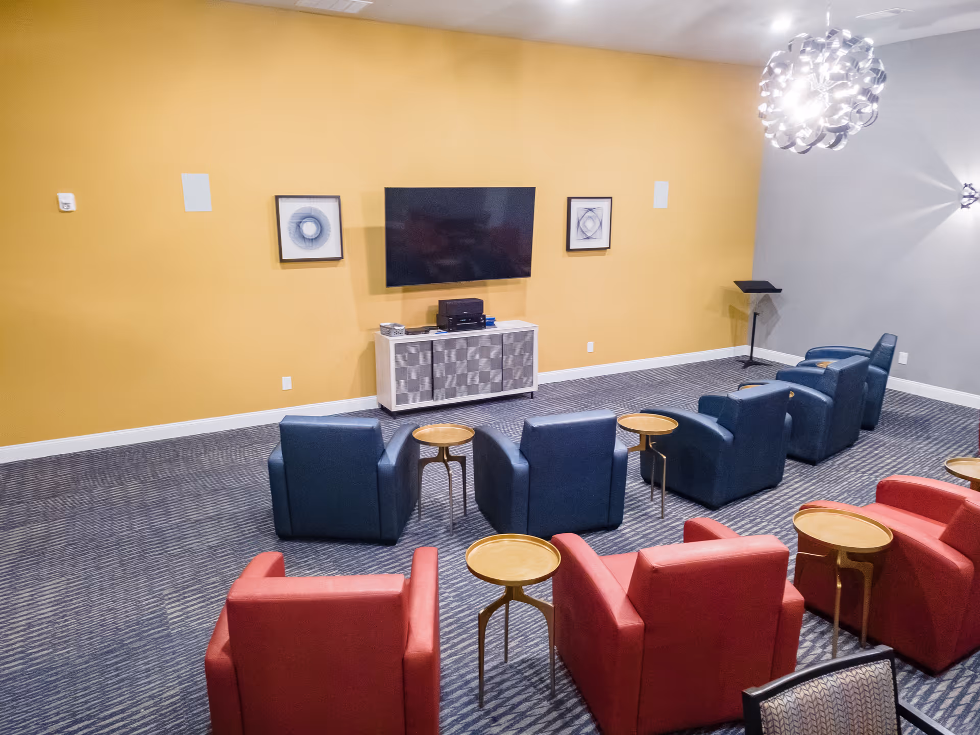 Community media room with a yellow accent wall, wall-mounted TV and rows of red and blue armchairs with small round side tables.