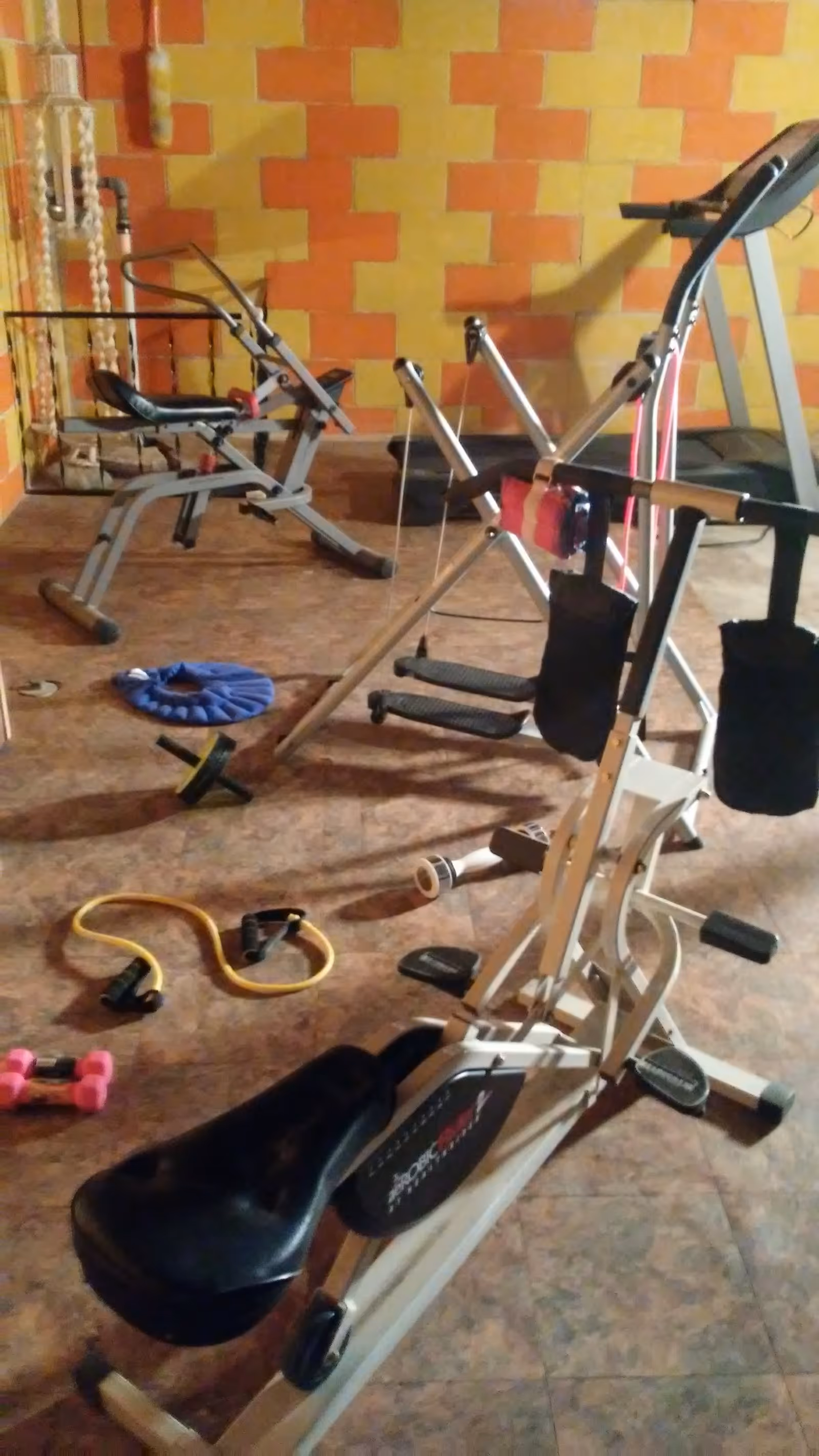 Small indoor exercise room with various workout equipment including a rowing machine, resistance bands, and dumbbells in front of a patterned wall.