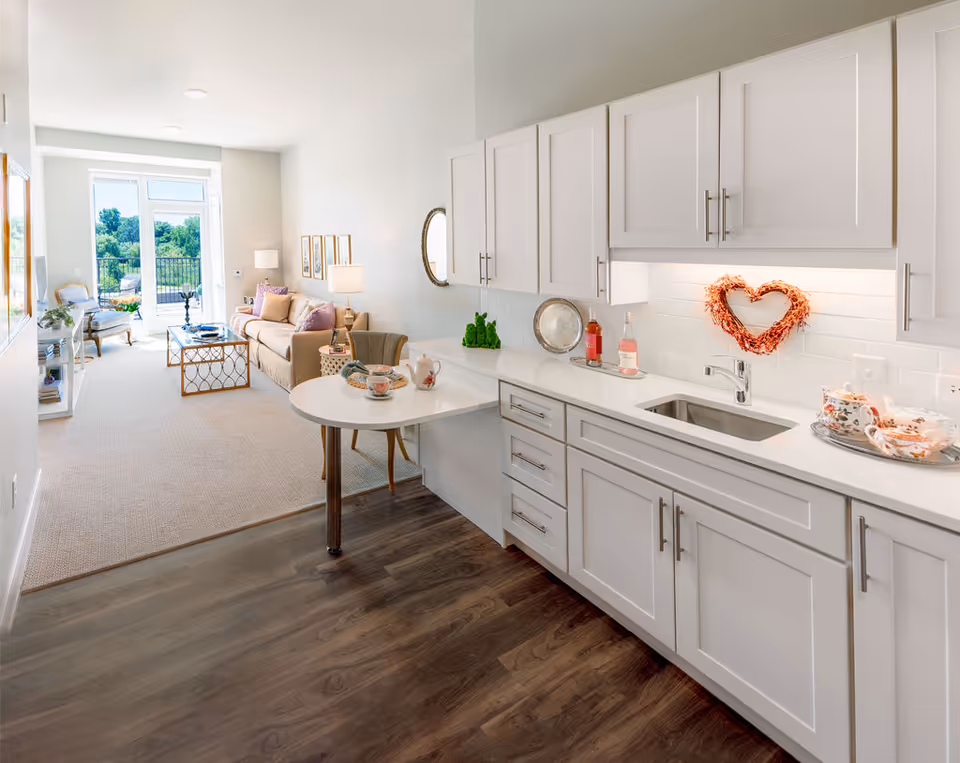 Bright open-plan kitchen with white cabinets and a sink opening into a sunlit living room with a sofa and balcony view.