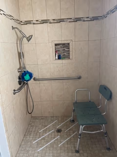 A tiled shower area with a wall-mounted showerhead, grab bars, a built-in niche holding toiletries, and a green shower chair with a backrest. The floor has a drain and anti-slip strips.
