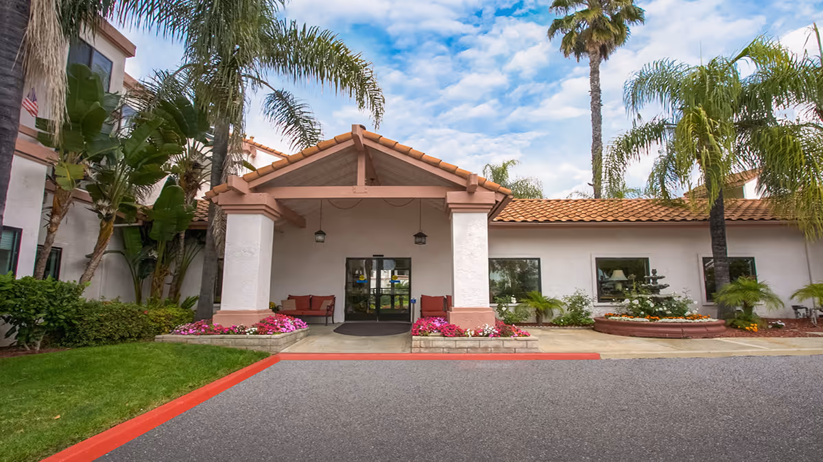 Front entrance of a senior living facility featuring a covered portico, seating, palm trees, flower beds and a driveway.