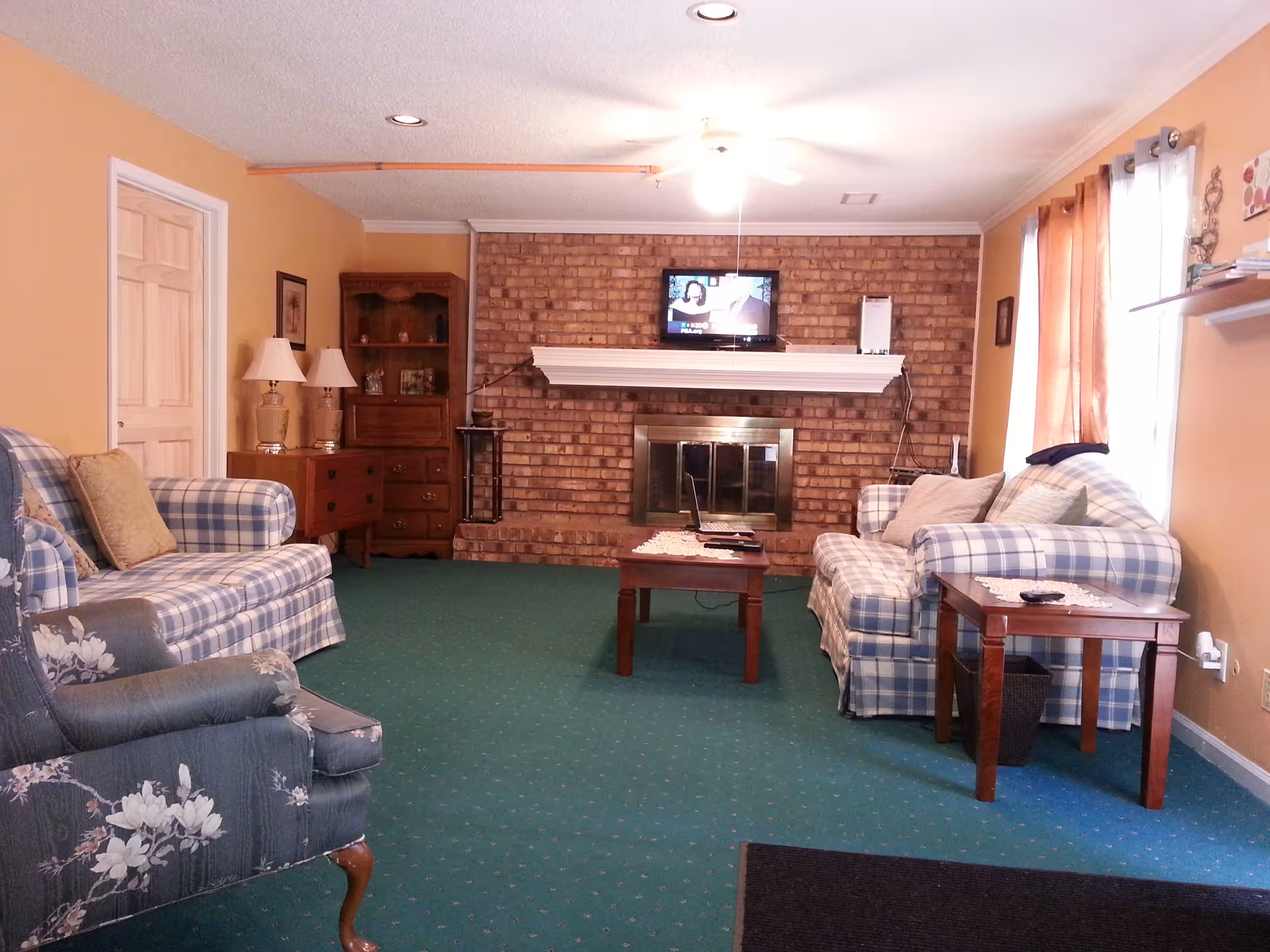 A cozy living room with green carpet, two plaid sofas with cushions, a floral armchair, a wooden coffee table, and side tables with lamps. A brick fireplace with a white mantel is centered on the far wall, above which a small television is mounted. The walls are painted a warm beige color, and there are windows with curtains on the right side.