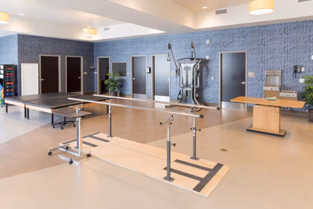 Spacious rehabilitation room with parallel walking bars, exercise equipment, and a ping-pong table against a blue-patterned wall.