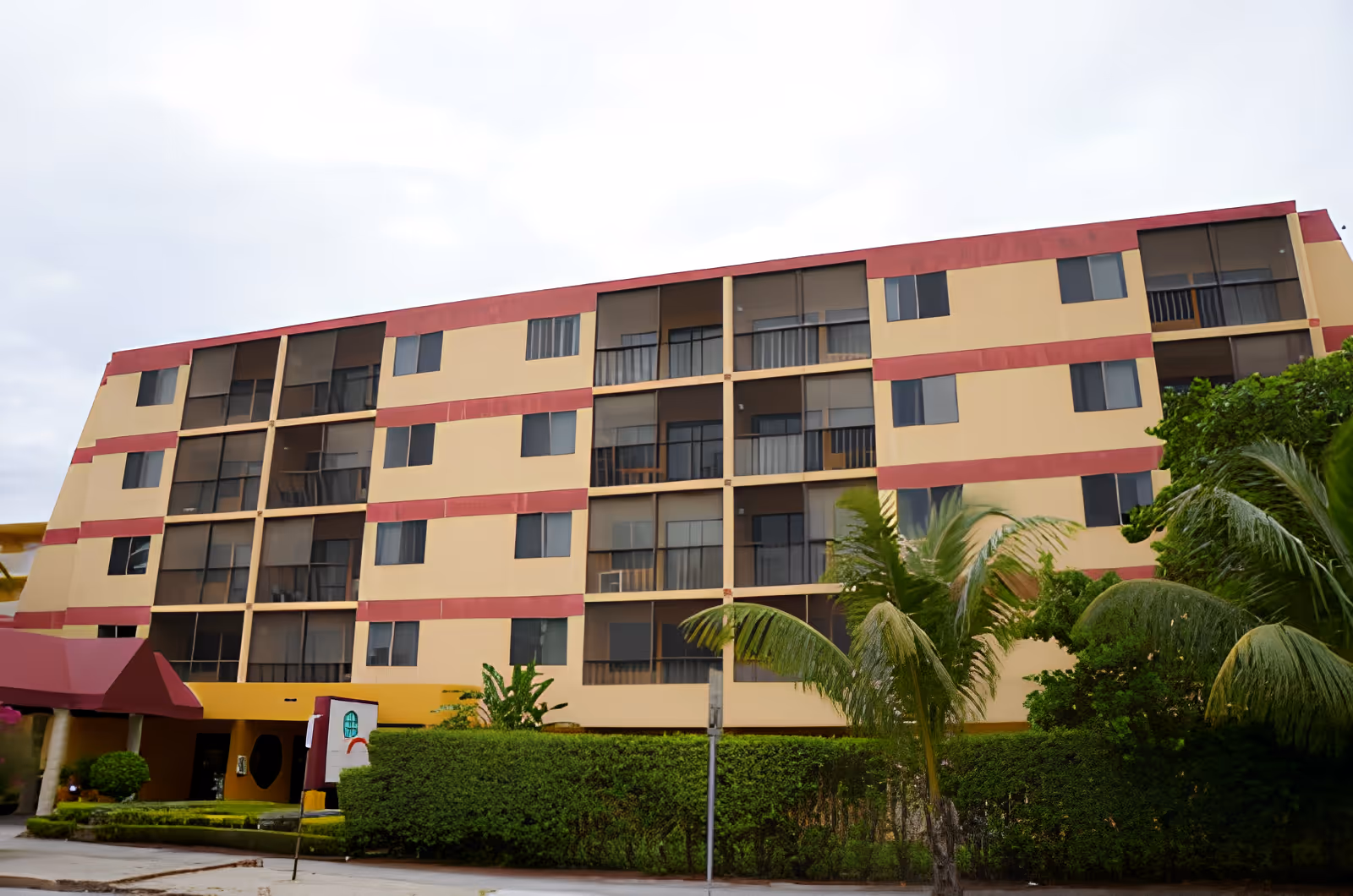 Exterior view of a four-story residential building with beige and red painted walls, multiple windows, and screened balconies. The building is surrounded by green bushes and palm trees under a cloudy sky.
