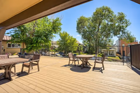 A sunlit wooden outdoor deck with round tables and chairs overlooking trees and a fenced yard.