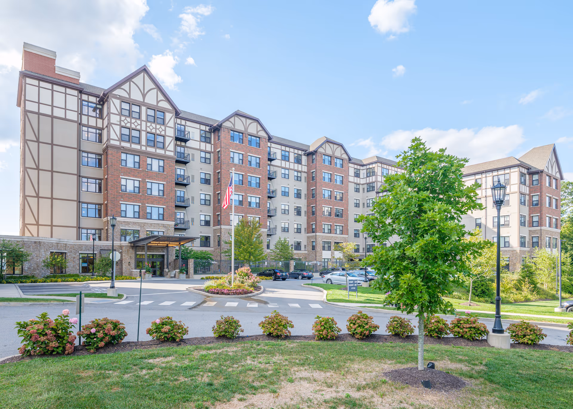 Front exterior of a multi-story senior living building with a circular driveway, landscaping, and an American flag.
