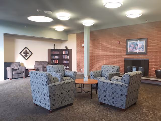 A communal lounge with four patterned armchairs arranged around a round table, additional seating, a bookshelf, and a fireplace against a brick wall.
