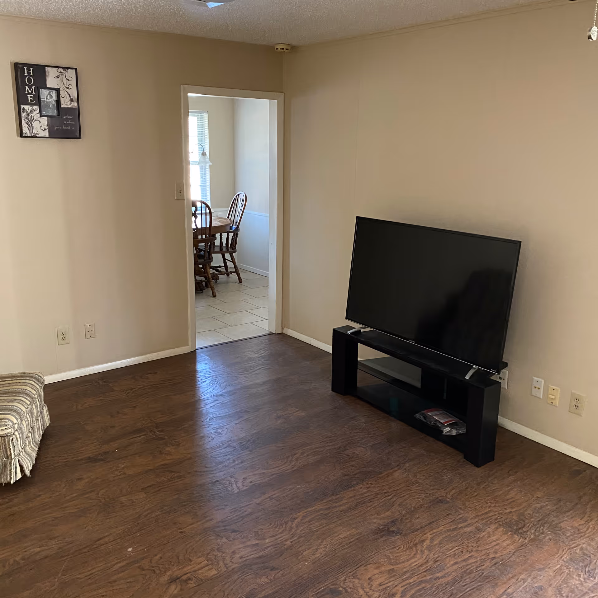A living room with wooden flooring, a flat-screen TV on a black TV stand, and a partial view of a striped upholstered chair. There is a doorway leading to a dining area with a wooden dining table and chairs, and a window with blinds. A decorative wall hanging with the word 'HOME' is visible on the left wall.
