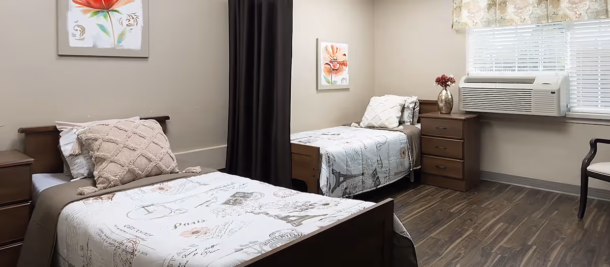A clean shared bedroom with two twin beds, wooden nightstands, wall art, and a window air conditioner.