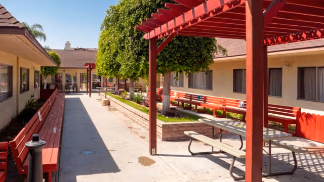 Outdoor courtyard area with red benches along the walls of single-story buildings, a picnic table under a red pergola, and neatly trimmed trees in raised brick planters.