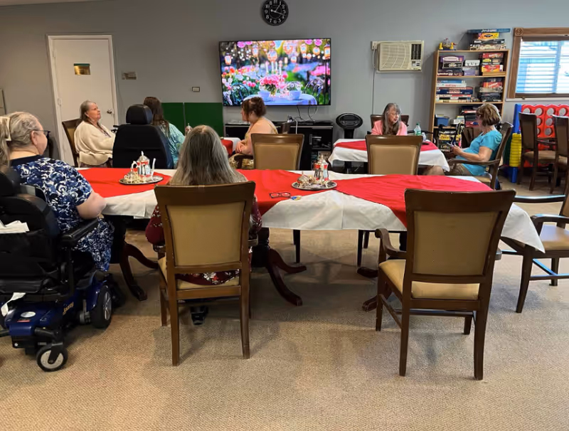 A group of elderly people sitting around tables covered with red and white tablecloths in a common room. They are engaged in conversation and watching a large television mounted on the wall displaying a colorful animated scene. The room has shelves with board games and an air conditioning unit on the wall.