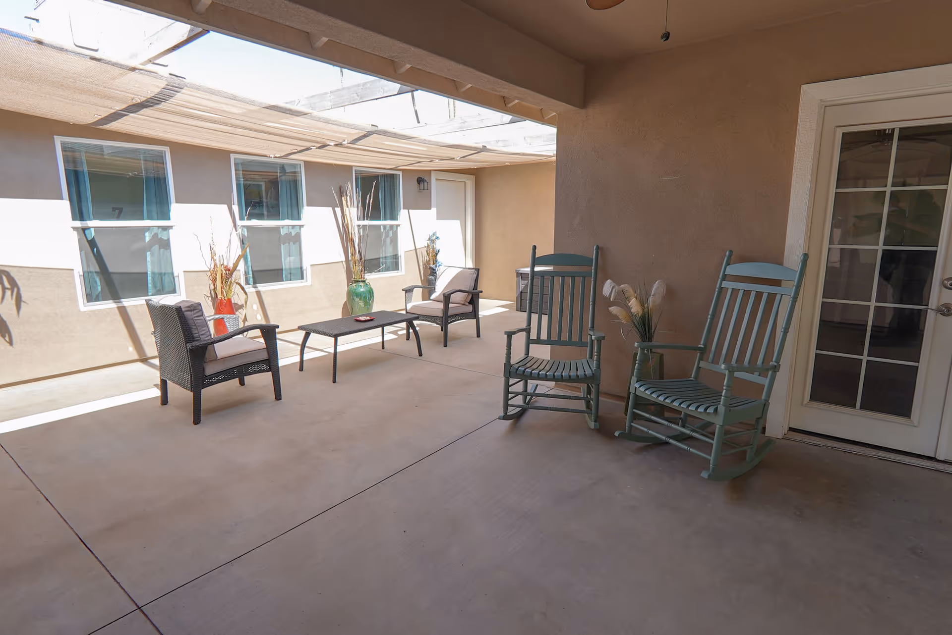 Covered outdoor patio area with two green rocking chairs, two cushioned armchairs, a black coffee table, and decorative vases with tall dried plants. The patio is adjacent to a building with windows and a glass door.