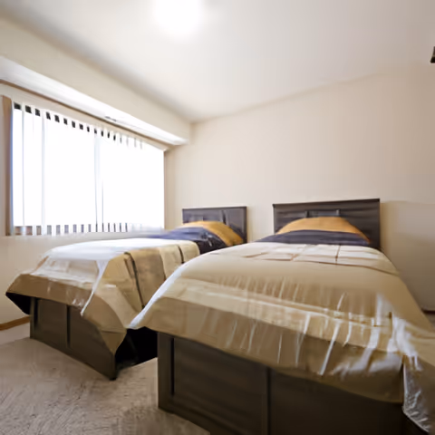 A bright bedroom with two single beds placed side by side, each with beige and brown bedding. The room has a large window with vertical blinds allowing natural light to fill the space. The walls are painted a light color and the floor is carpeted.