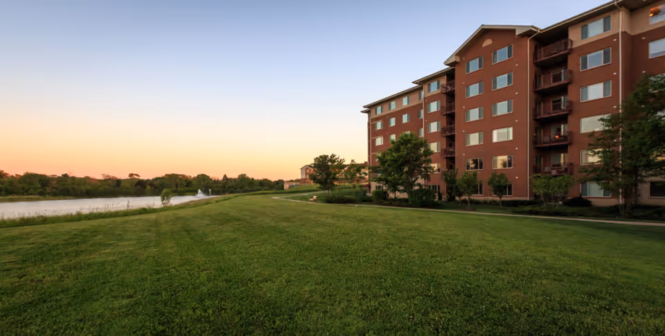 A large multi-story brick building with balconies overlooking a well-maintained grassy lawn and a pond with a fountain, under a clear sky at sunset.