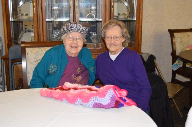 Two elderly women sitting at a round table in a room with a china cabinet in the background. One woman is wearing a patterned hat, glasses, a teal jacket, and a purple shirt, smiling warmly. The other woman is wearing glasses and a purple sweater, also smiling. A pink and white crocheted blanket is spread out on the table in front of them.