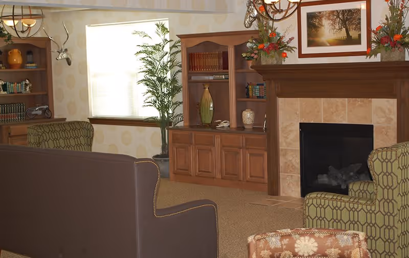 A cozy living room area with a brown sofa, patterned armchairs, a wooden bookshelf filled with books and decorative items, a fireplace with a wooden mantel adorned with flower arrangements and a framed picture above it, and a window with blinds partially open letting in natural light.