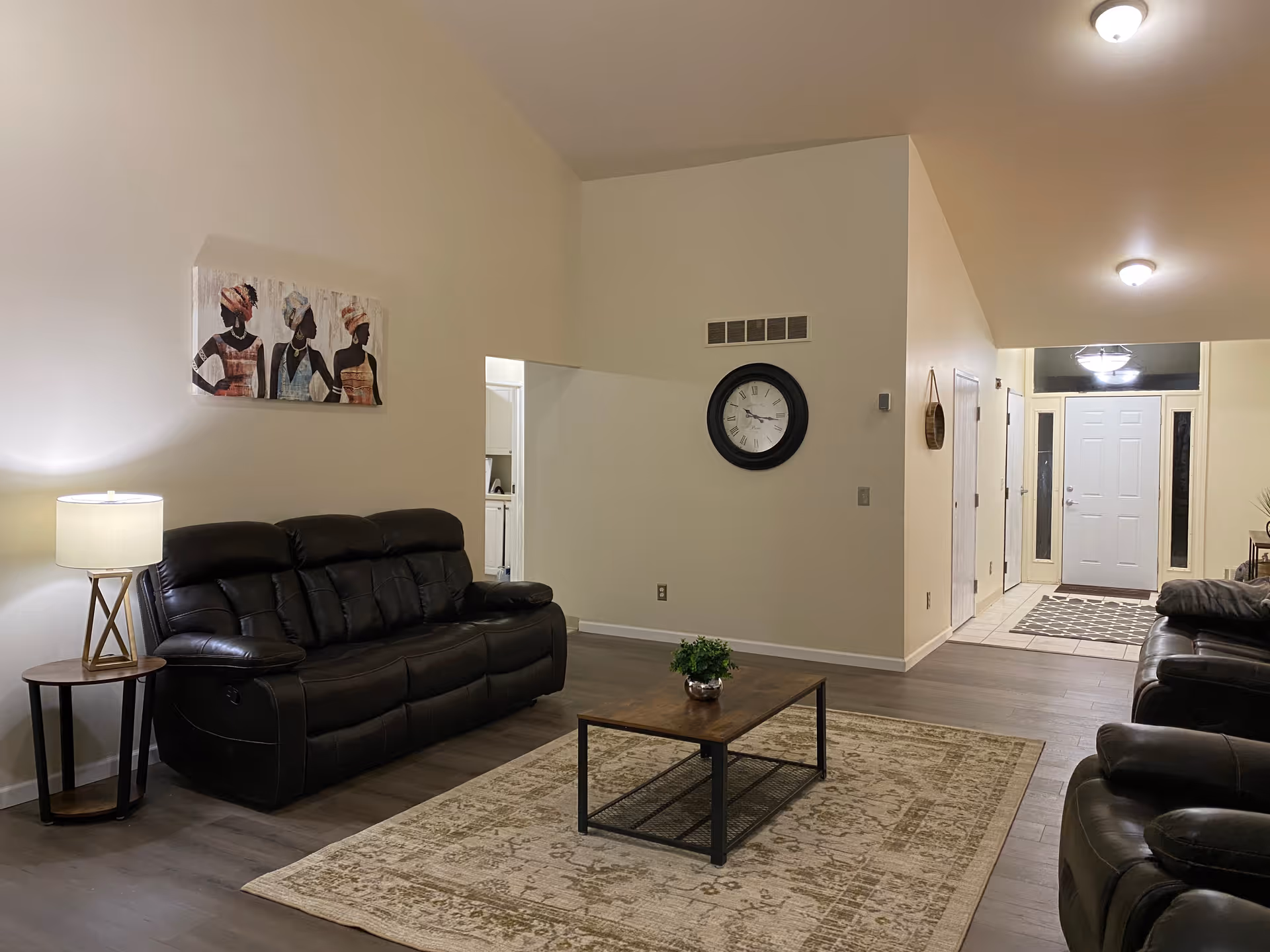 A cozy living room with two dark leather sofas facing each other, a wooden coffee table with a small plant on it, a patterned area rug, a side table with a lamp, a large wall clock, and a painting of three women on the wall. The room has light-colored walls and wood flooring, with a hallway leading to a white front door.