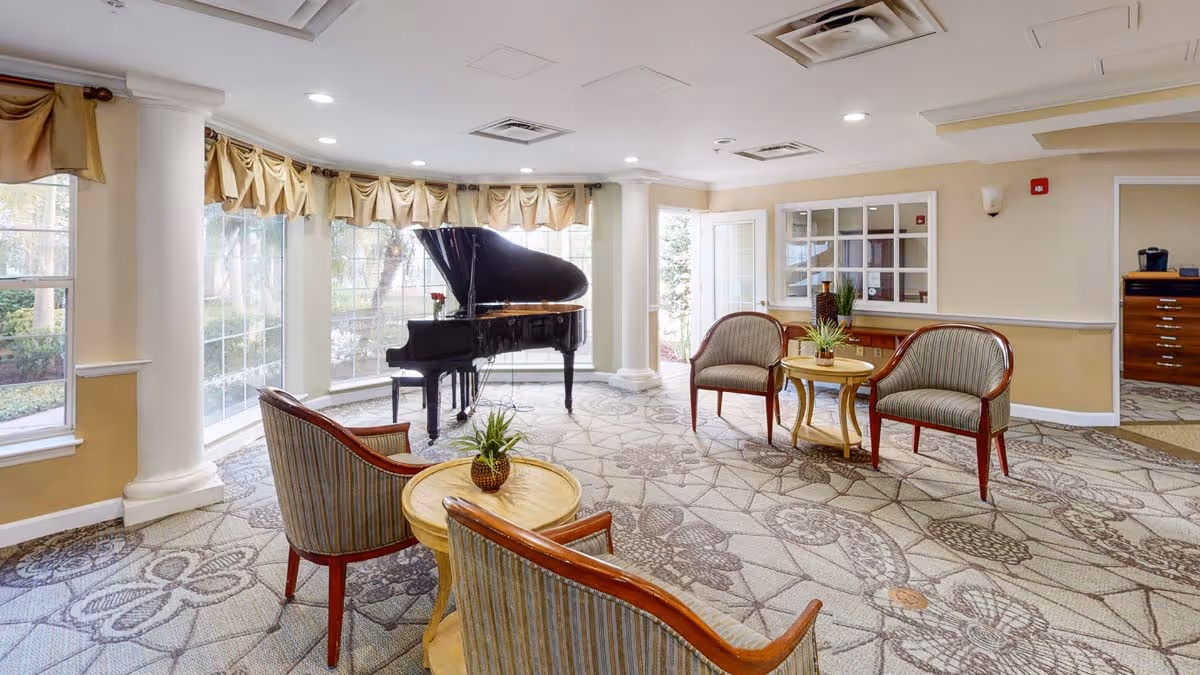 Bright senior living lounge with a grand piano by large windows and grouped seating around small tables.
