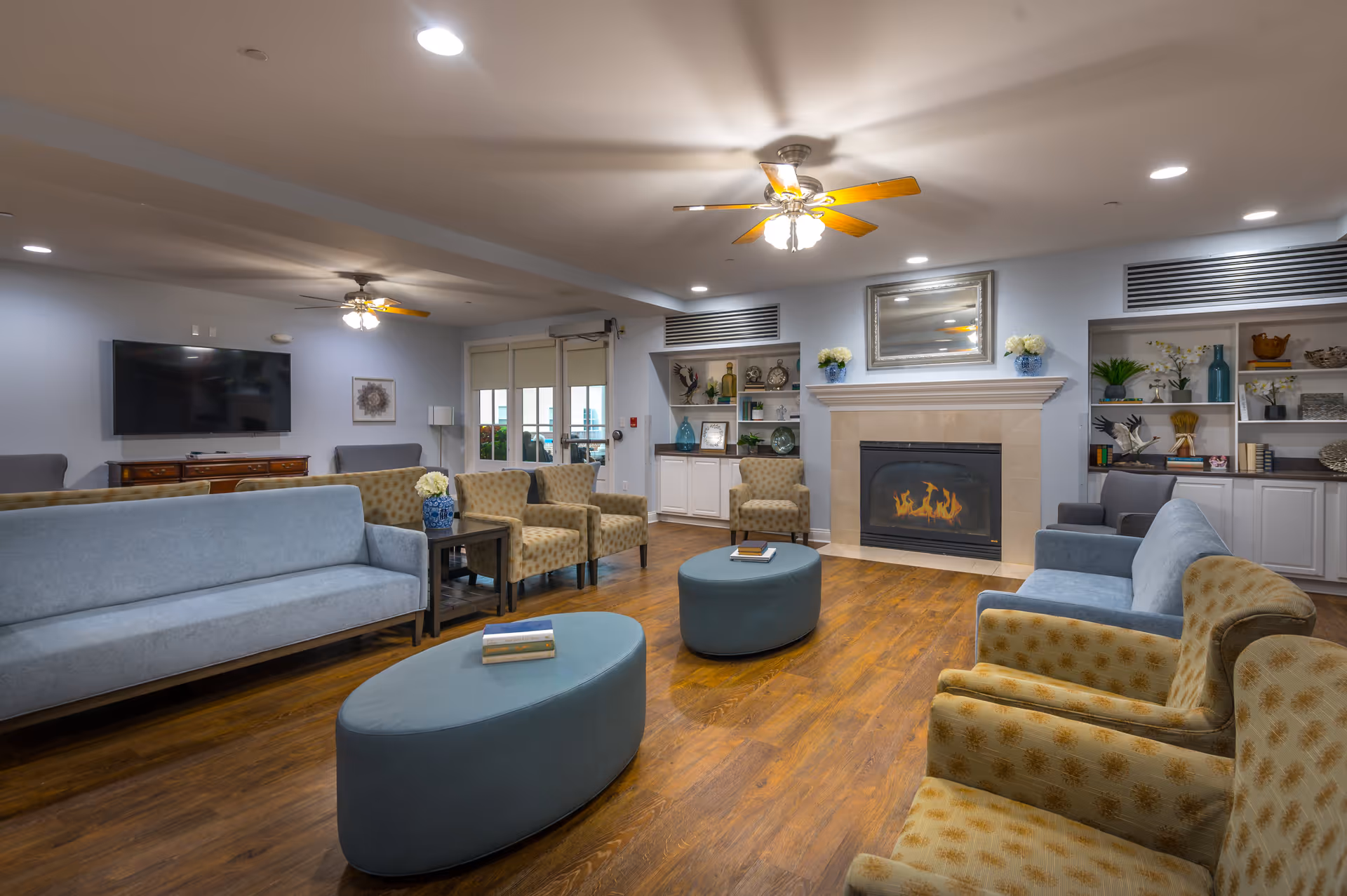 Well-lit communal living room with sofas, armchairs, ottomans, a fireplace, built-in shelving, and a wall-mounted TV.