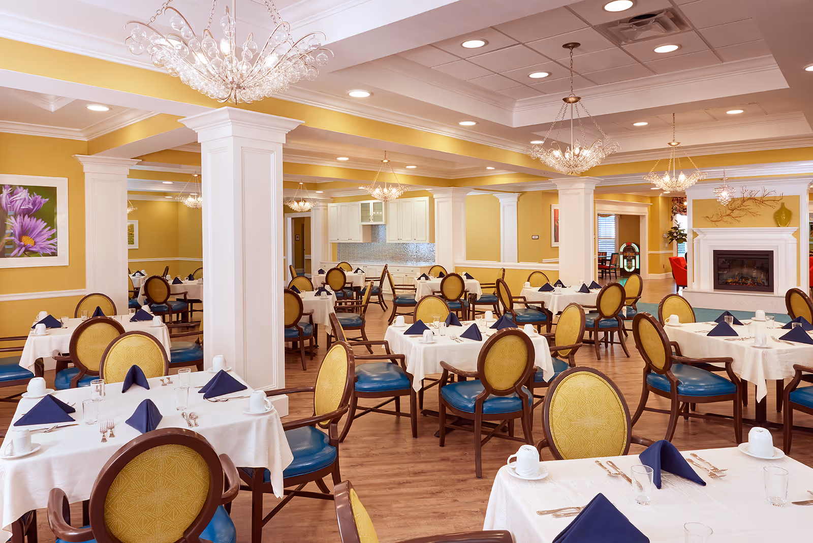 A spacious and elegant dining room with multiple round tables covered in white tablecloths, each set with blue folded napkins, cups, glasses, and silverware. The room features yellow walls with white trim, wooden flooring, decorative columns, chandeliers, and a fireplace with a mantel decorated with a green vase and branches. There is framed floral artwork on the walls and a jukebox visible in the background.
