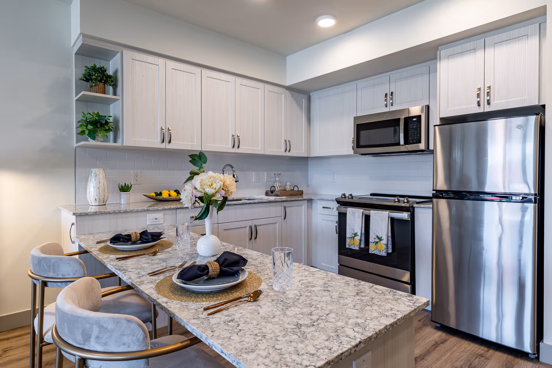 Modern kitchen with light-colored cabinets, stainless steel refrigerator, stove, and microwave. A marble countertop island with two place settings, each with a plate, napkin, and glass. Decorative plants and flowers are placed on the countertop and shelves.