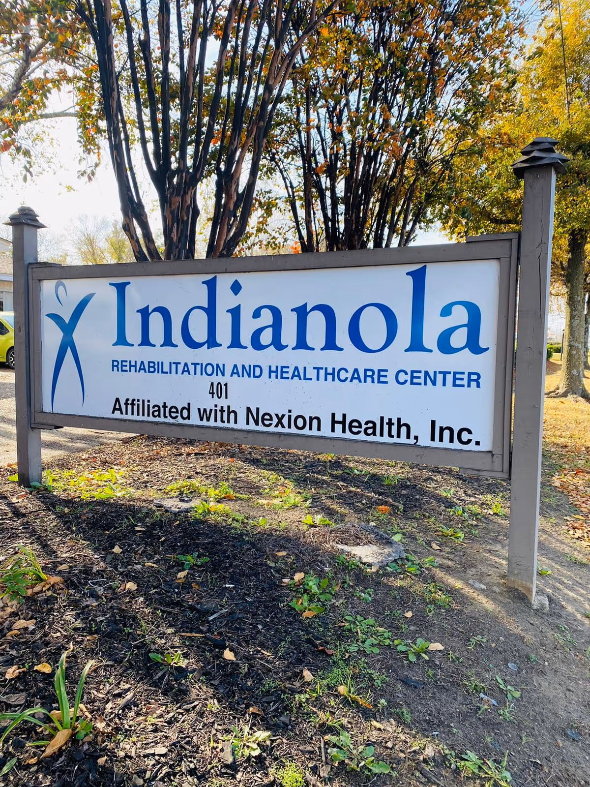 Outdoor sign for Indianola Rehabilitation and Healthcare Center, affiliated with Nexion Health, Inc., surrounded by trees and plants with sunlight filtering through.