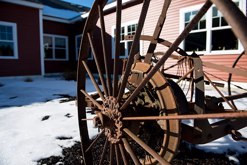 Rusty vintage wagon wheel sculpture in foreground with snow and red building exterior.