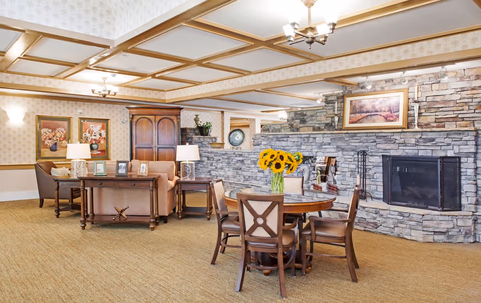 A cozy living room area in a senior living facility featuring a stone fireplace with a framed painting above it. In front of the fireplace is a round wooden table with four chairs and a vase of sunflowers. To the left, there is a wooden cabinet, a sofa, two table lamps, and framed floral artwork on the wall. The room has a beige carpet and a ceiling with wooden beams and multiple light fixtures.