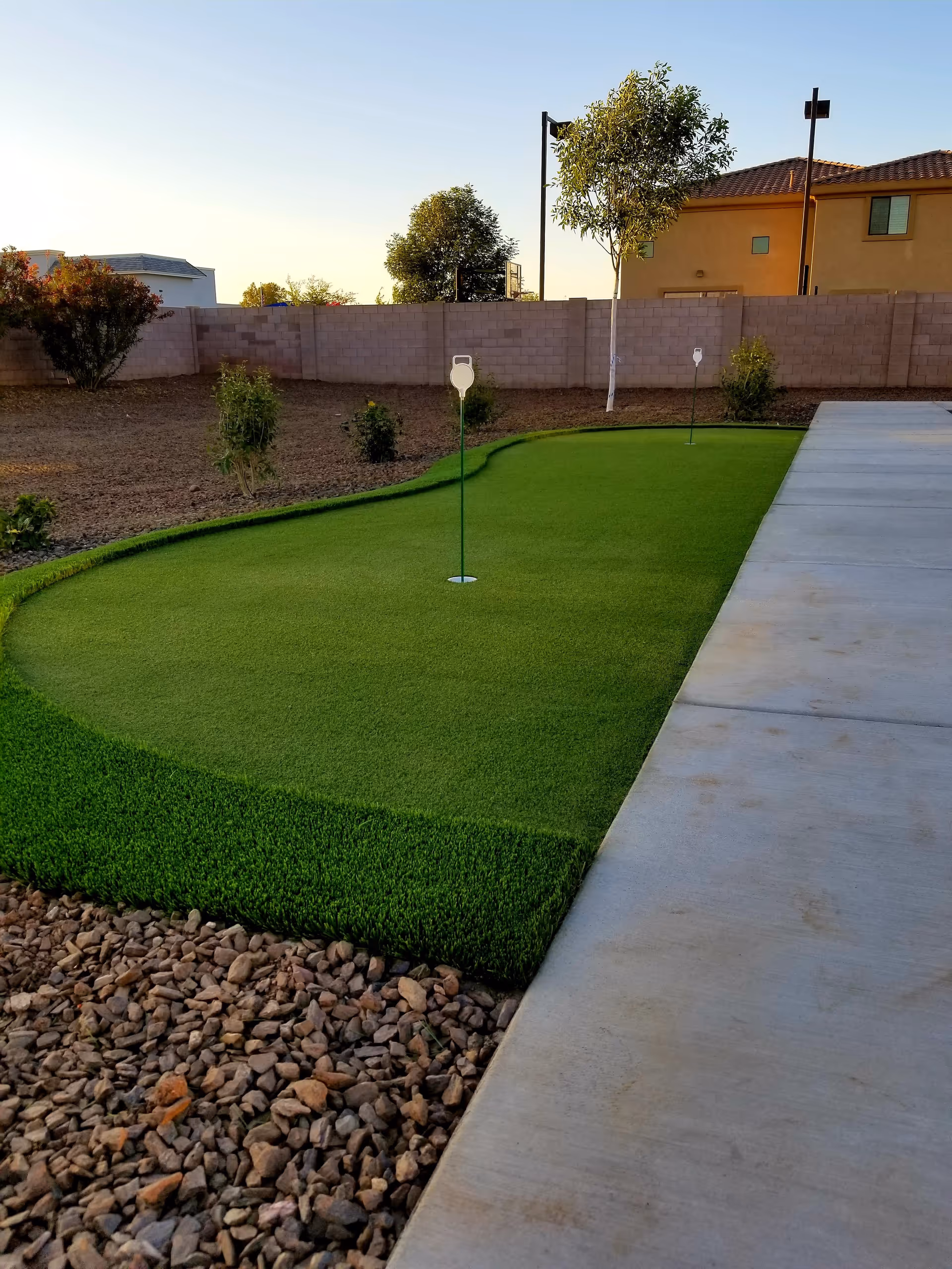 Outdoor putting green with two golf holes and flags, surrounded by a gravel area and a concrete walkway. There are small bushes and trees along a brick wall fence, with a house and clear sky in the background.