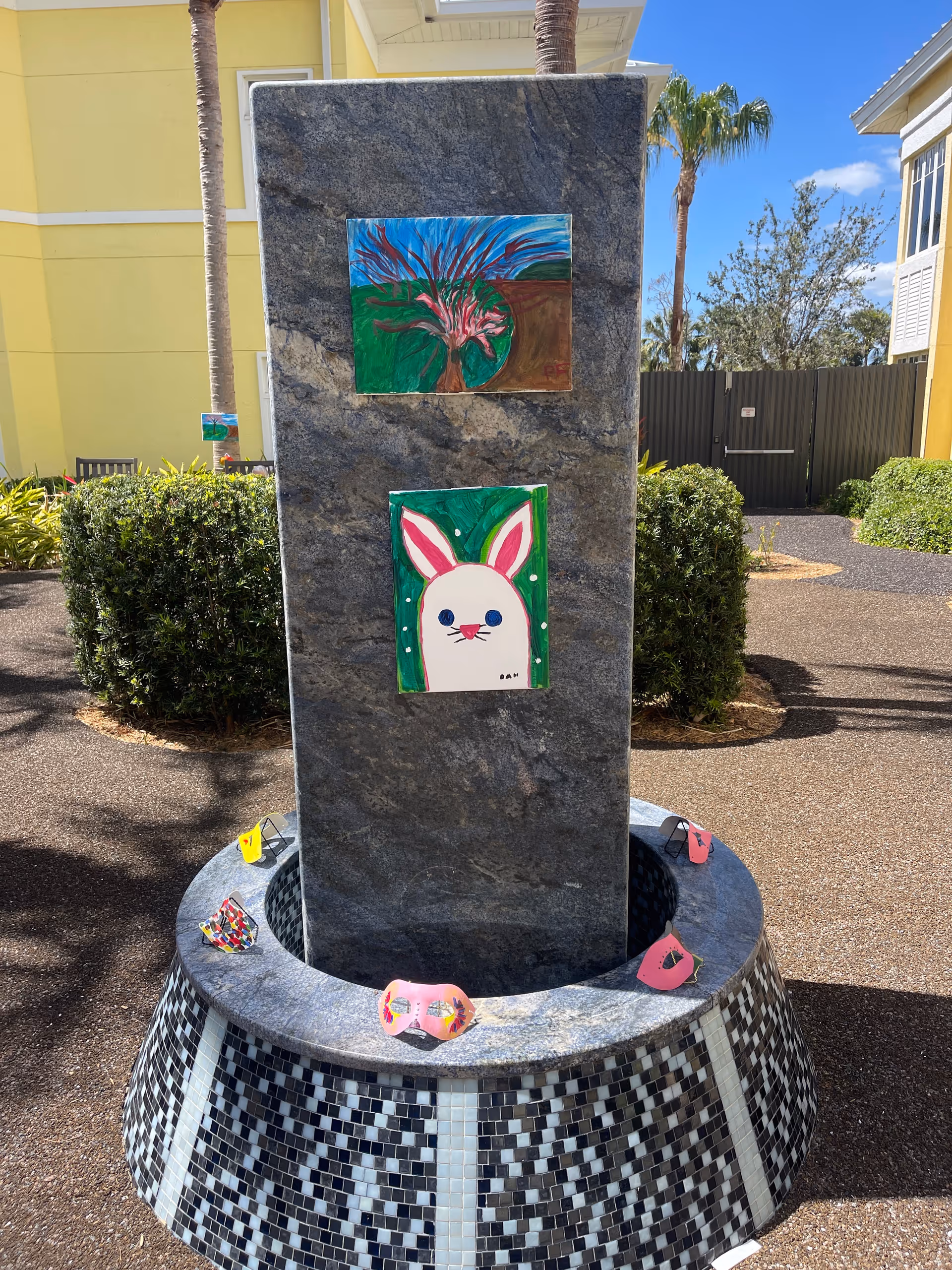Outdoor stone pillar with two colorful paintings attached, one of a tree and one of a white bunny with pink ears. The pillar is surrounded by a circular tiled base with several decorated face masks placed on it. The background shows bushes, palm trees, a yellow building, and a clear blue sky.