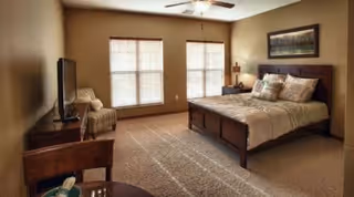 A bedroom with a large wooden bed with pillows and a comforter, two windows with blinds letting in natural light, a ceiling fan, a TV on a wooden stand, and a chair in the corner.