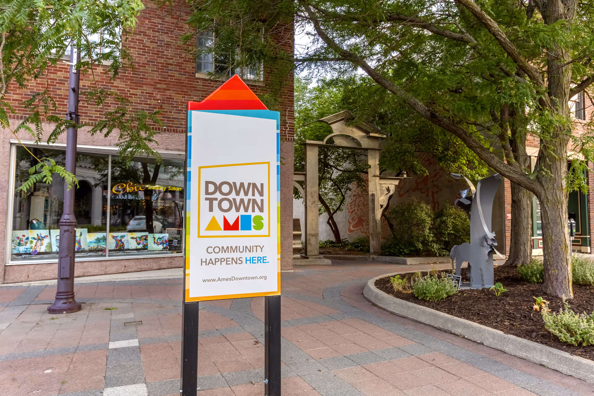 Outdoor view of a downtown area with a sign that reads 'DOWNTOWN AMES COMMUNITY HAPPENS HERE. www.AmesDowntown.org'. The scene includes a brick building with a storefront window displaying colorful artwork, a lamppost, trees, a small landscaped area with plants, and a metal sculpture of an animal.