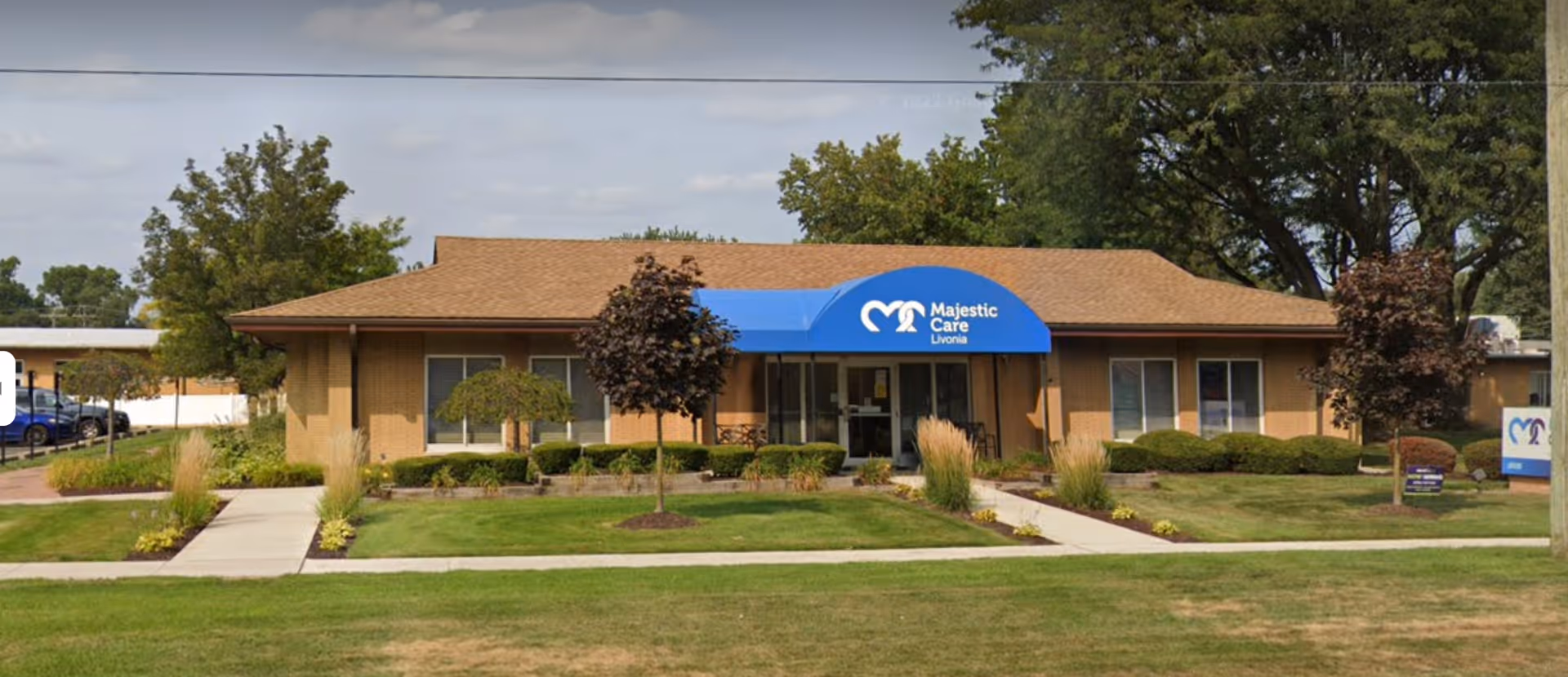 Single-story brick building with a brown roof and a blue awning over the entrance that reads 'Majestic Care Livonia'. The building is surrounded by green grass, small trees, and shrubs, with a sidewalk leading to the entrance.