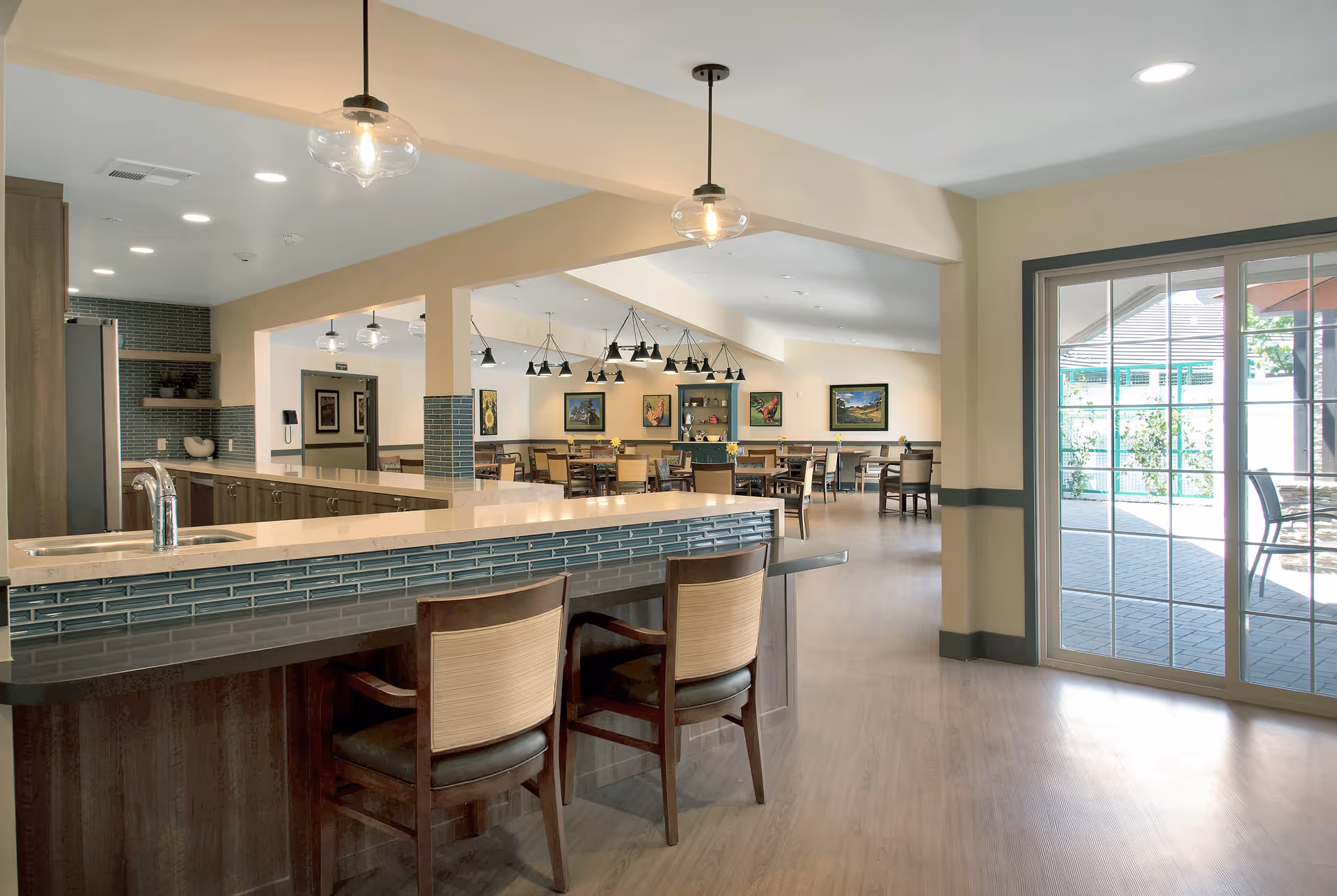 A bright and spacious dining area in an assisted living facility featuring a kitchen counter with two chairs, pendant lights hanging from the ceiling, multiple dining tables and chairs, framed artwork on the walls, and large sliding glass doors leading to an outdoor patio.