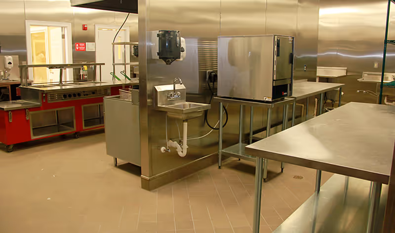 Commercial stainless-steel kitchen with prep tables, sinks, and cooking equipment.