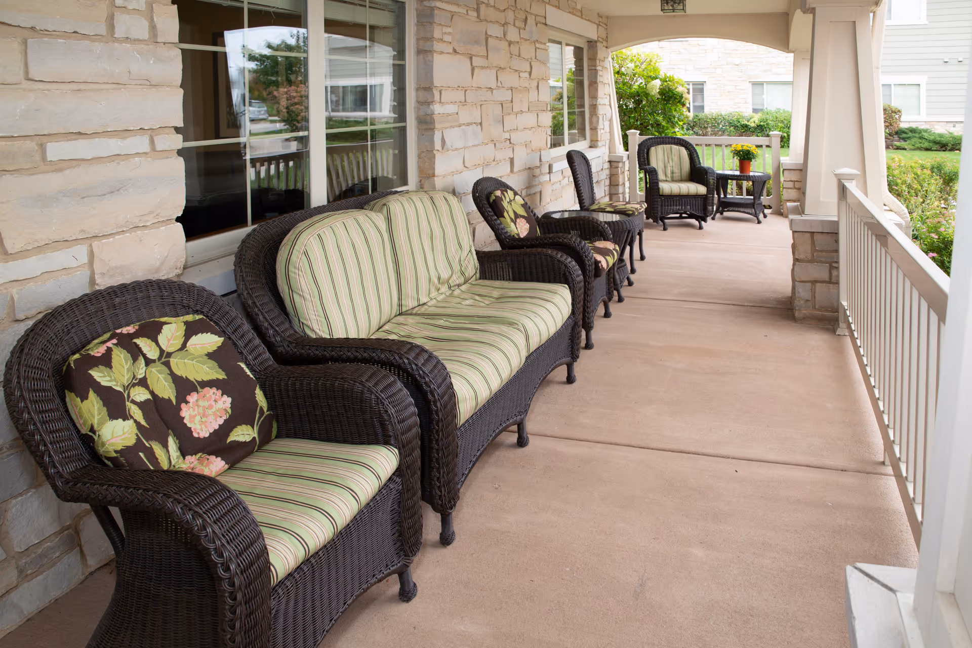 Covered outdoor porch area with wicker furniture including chairs and a loveseat with striped and floral cushions, stone wall, and railing overlooking a garden.