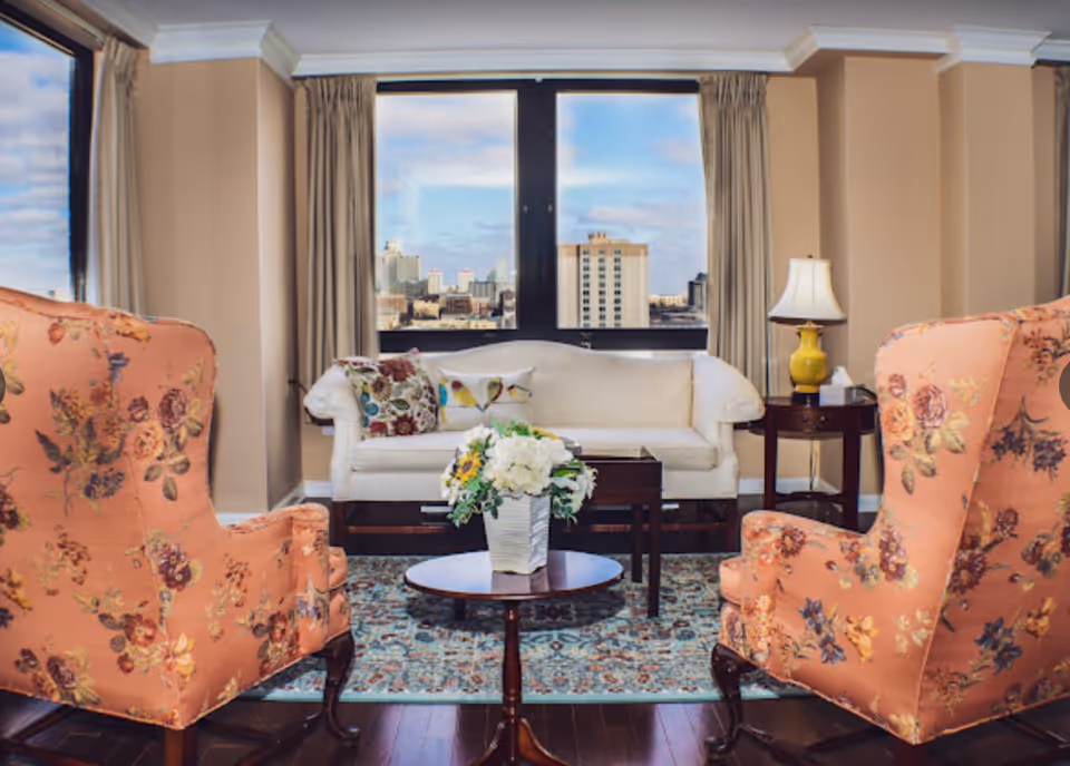 A cozy living room with two floral upholstered armchairs facing a white sofa adorned with decorative pillows. A small round wooden table with a flower arrangement is in the center on a patterned rug. Behind the sofa, large windows reveal a cityscape view under a partly cloudy sky. A side table with a yellow lamp and tissue box is next to the sofa.