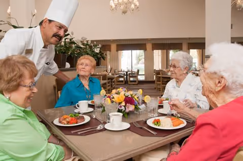 A chef speaks with four elderly women seated around a dining table set with plates, cups, and a floral centerpiece in a communal dining room.