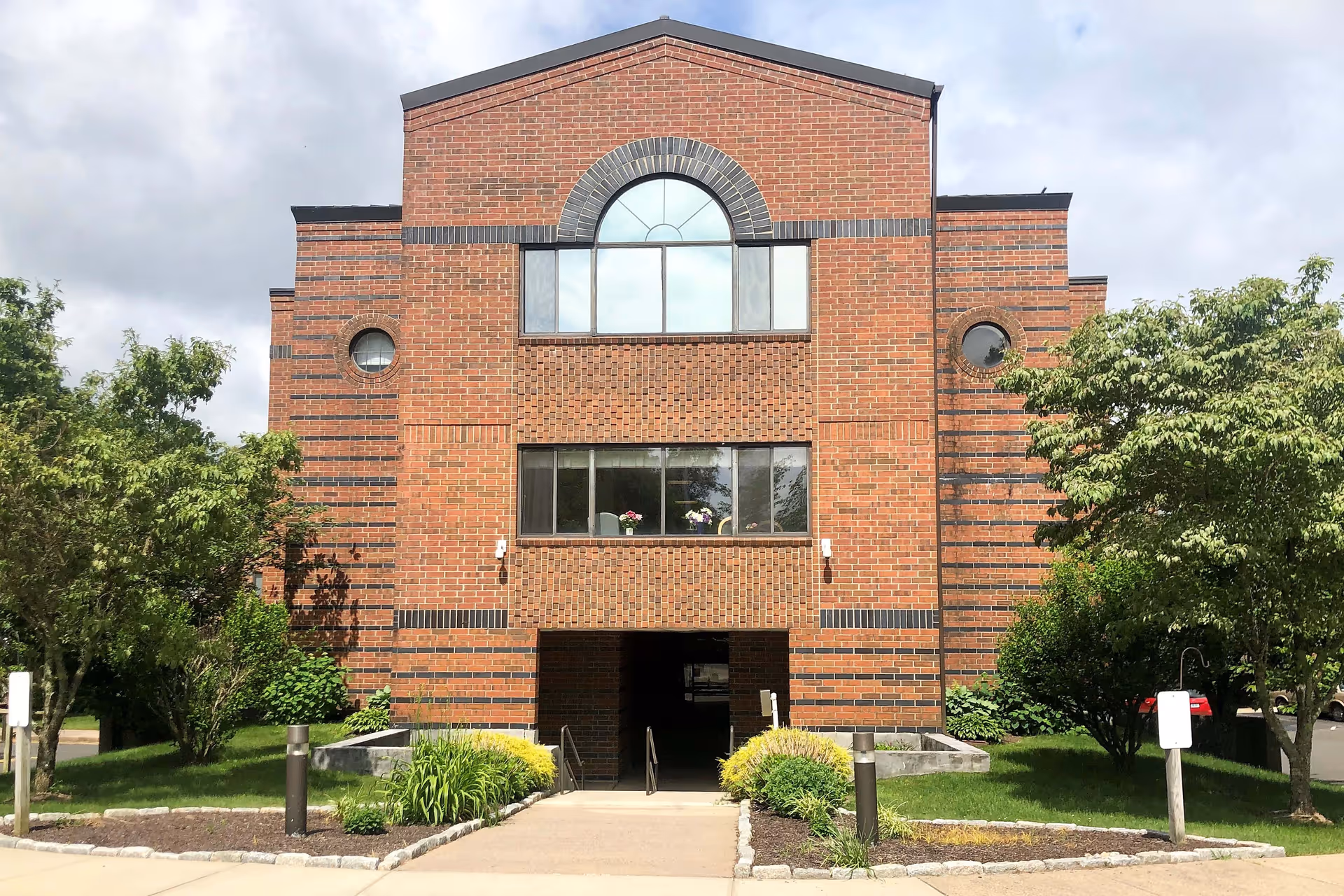 Front exterior view of a three-story brick building with large windows and an arched window at the top center. The building is surrounded by green trees and shrubs, with a paved walkway leading to the entrance.