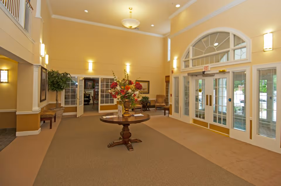 Spacious senior living lobby with a round table topped by a floral arrangement, seating areas, and glass double doors at the entrance.